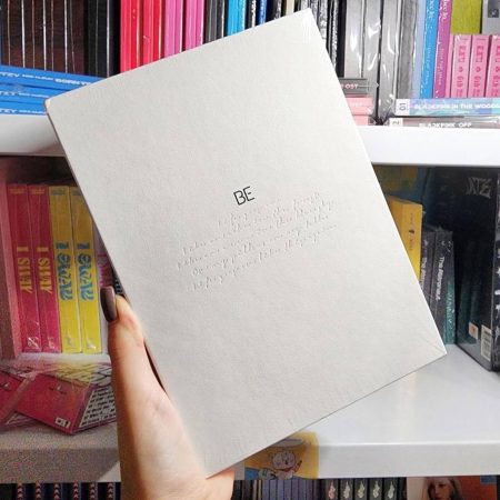 Альбом BTS BE (Essential Edition) у світло-сірій обкладинці з тисненням, на тлі полиць з K-pop альбомами.