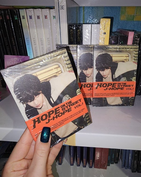 Рука тримає альбом j-hope «Hope on the Street Vol.1», ще один примірник на полиці з альбомами BTS на задньому плані