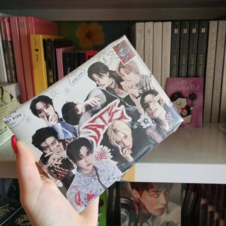 Рука тримає альбом Stray Kids на фоні полиці з K-pop альбомами, включно з BTS Love Yourself
