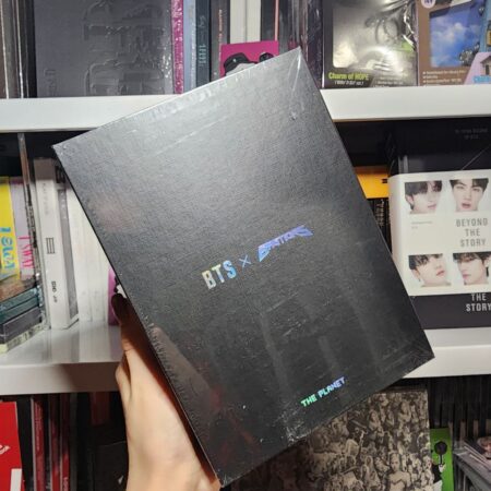 Рука тримає чорну коробку BTS x Bastons — The Planet на фоні полиці з K-pop мерчем