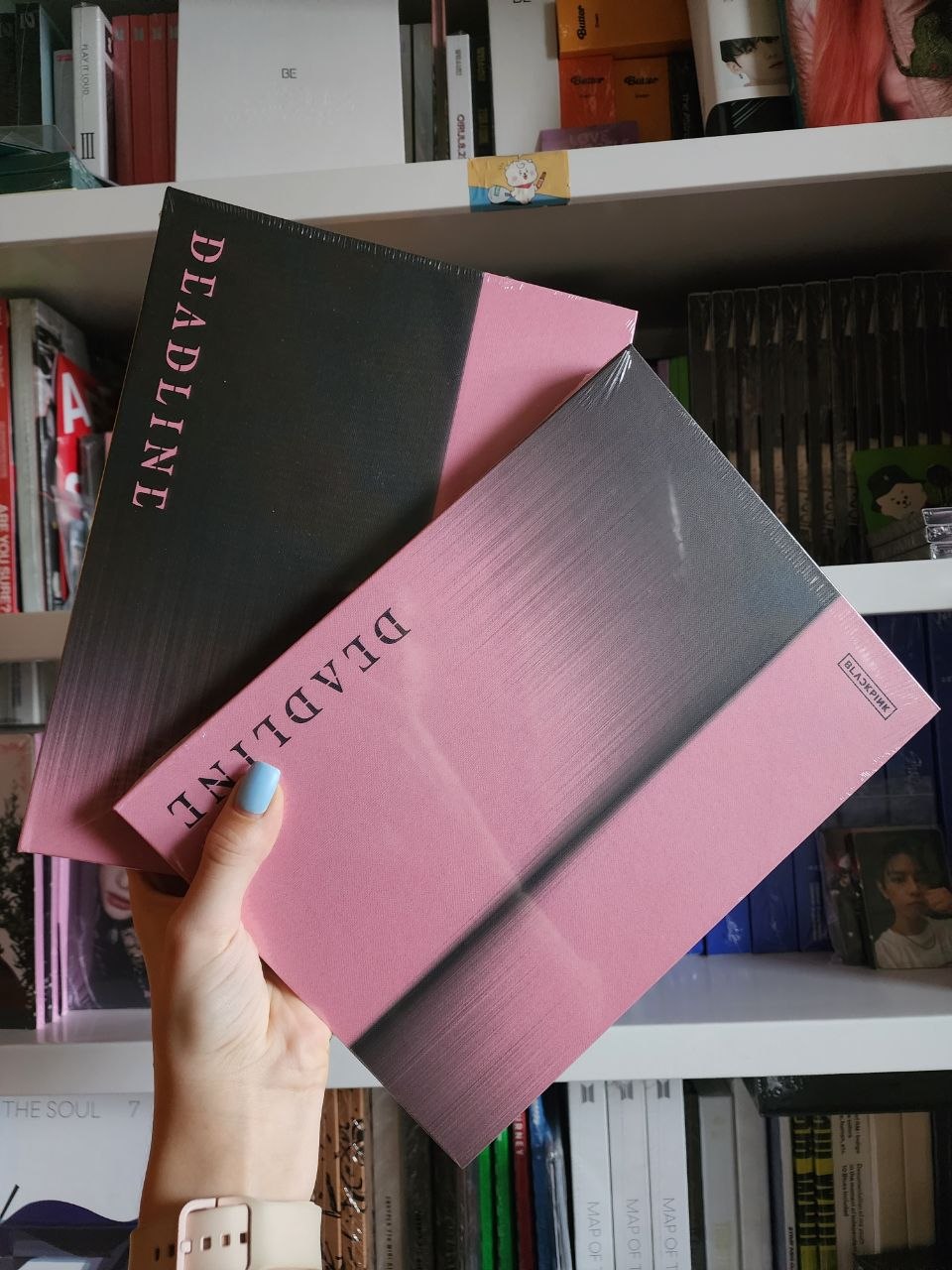 Рука тримає два альбоми BLACKPINK «DEADLINE» у чорній та рожевій версіях на фоні полиць з K-pop альбомами