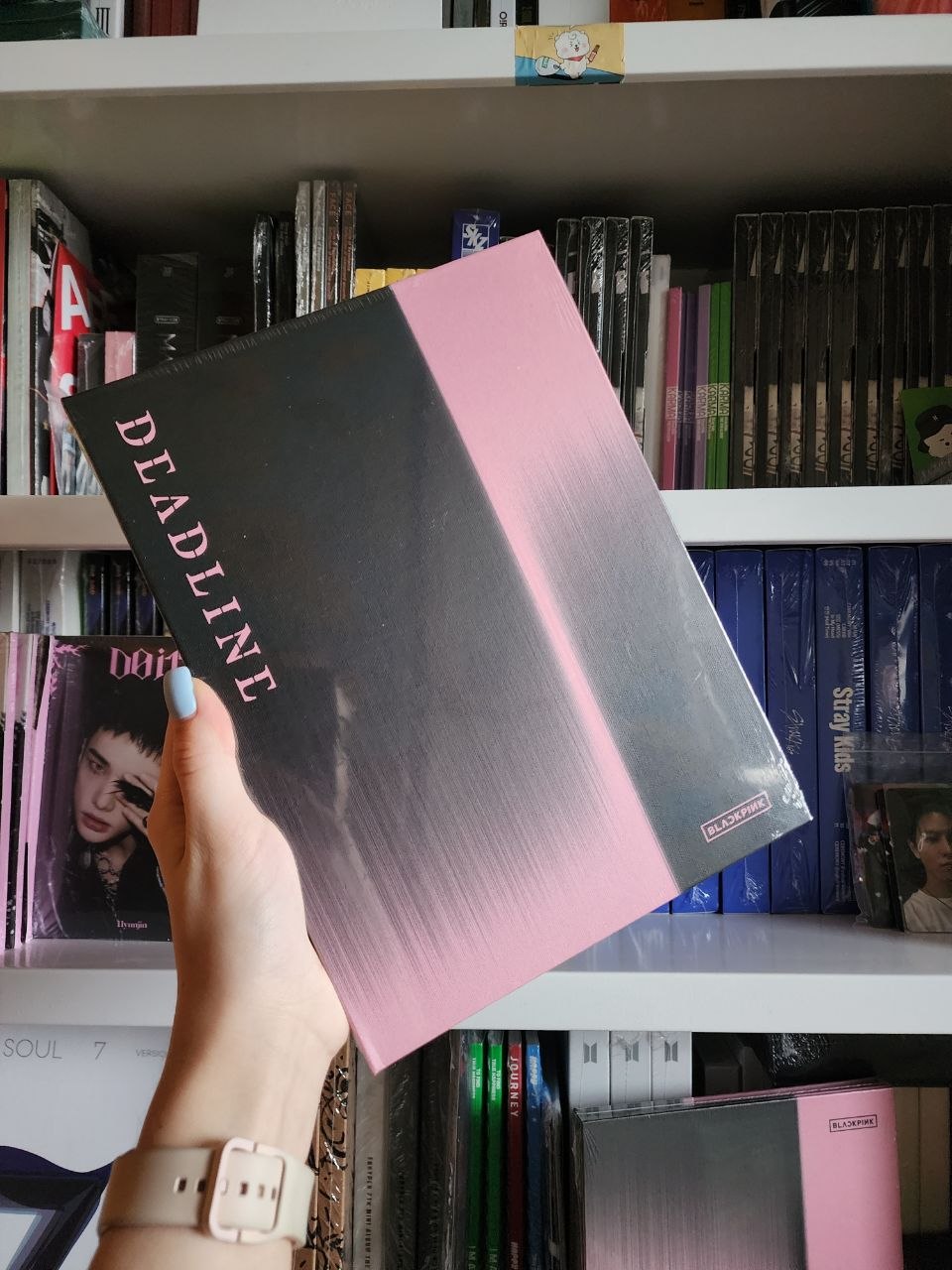Рука тримає альбом BLACKPINK «Deadline» у чорно-рожевій обкладинці на фоні полиць з K-pop альбомами